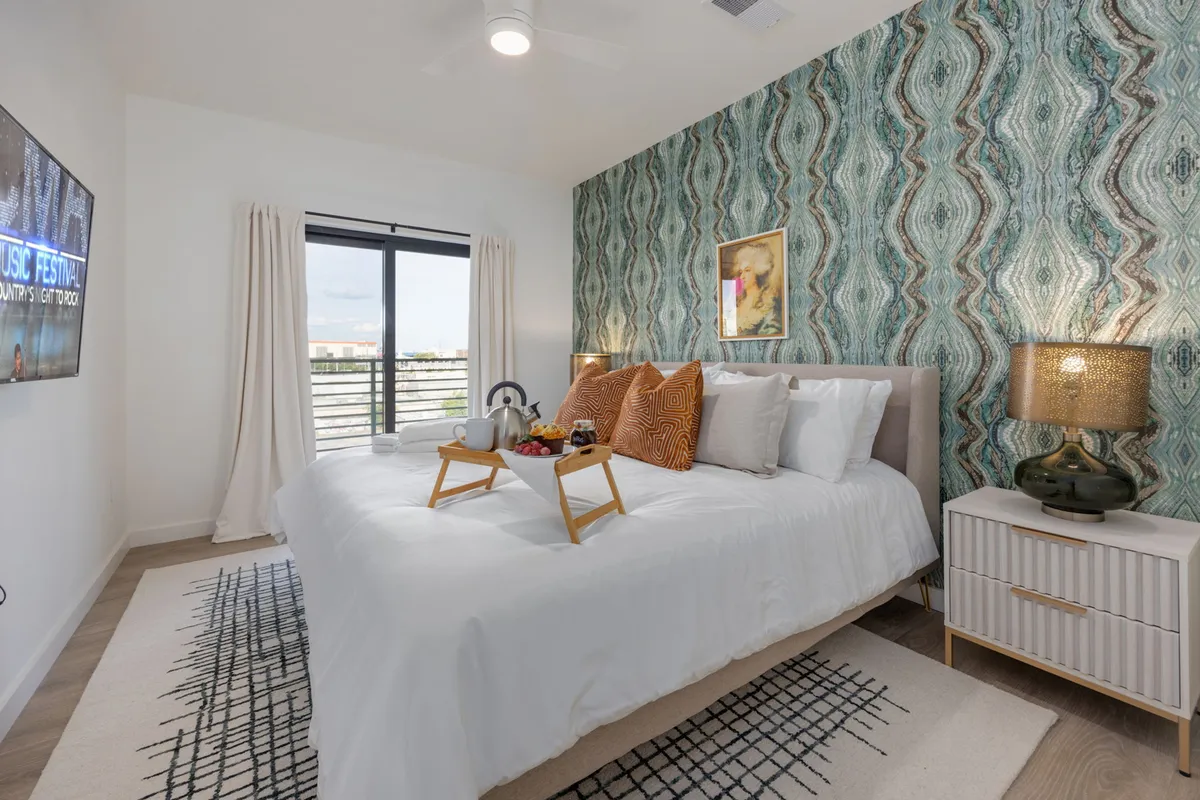 This image features a large bed adorned with decorative pillows, a textured accent wall, a nightstand with a lamp, and a TV mounted on the wall. The setting is both stylish and cozy, evoking a restful atmosphere ideal for unwinding after a long day.