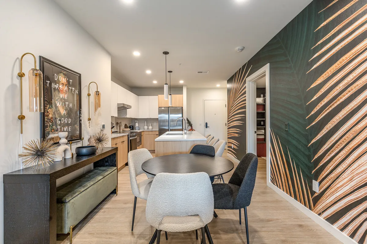 This interior shot centers on a dining table adjacent to a kitchen area, with a feature wall of lush palm leaf art and a framed quote. The mood is contemporary and relaxed, ideal for meals, gatherings, or socializing.