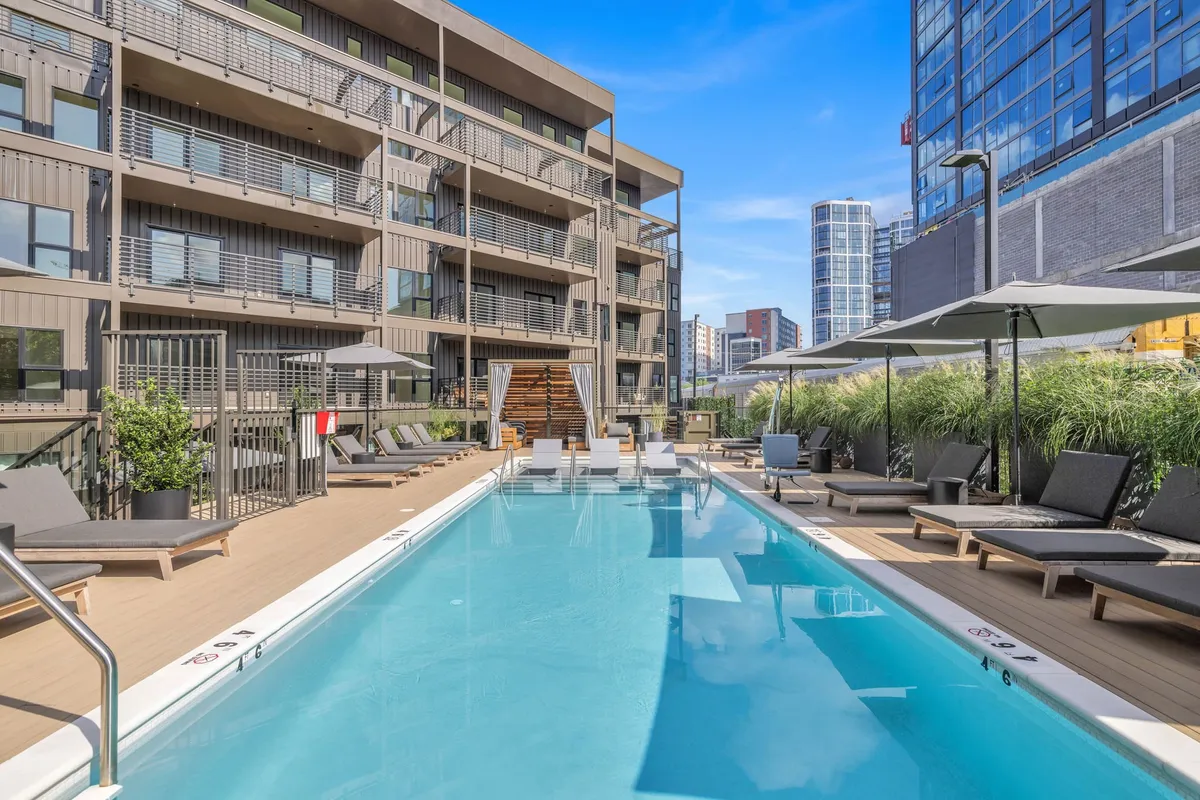 An inviting swimming pool with crystal-clear water, surrounded by lounge chairs and umbrellas. The building's modern facade is visible in the background, setting the stage for a resort-like experience. The overall atmosphere is relaxed, with ample space for sunbathing, swimming, or socializing.
