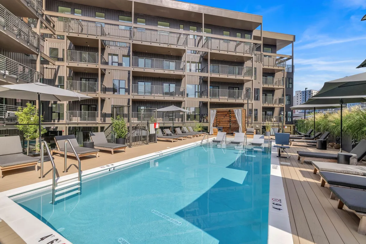 A beautifully maintained outdoor swimming pool surrounded by lounge chairs, umbrellas, green landscaping, and the building's facade. The area is spacious and inviting, ideal for both relaxation and social gatherings.