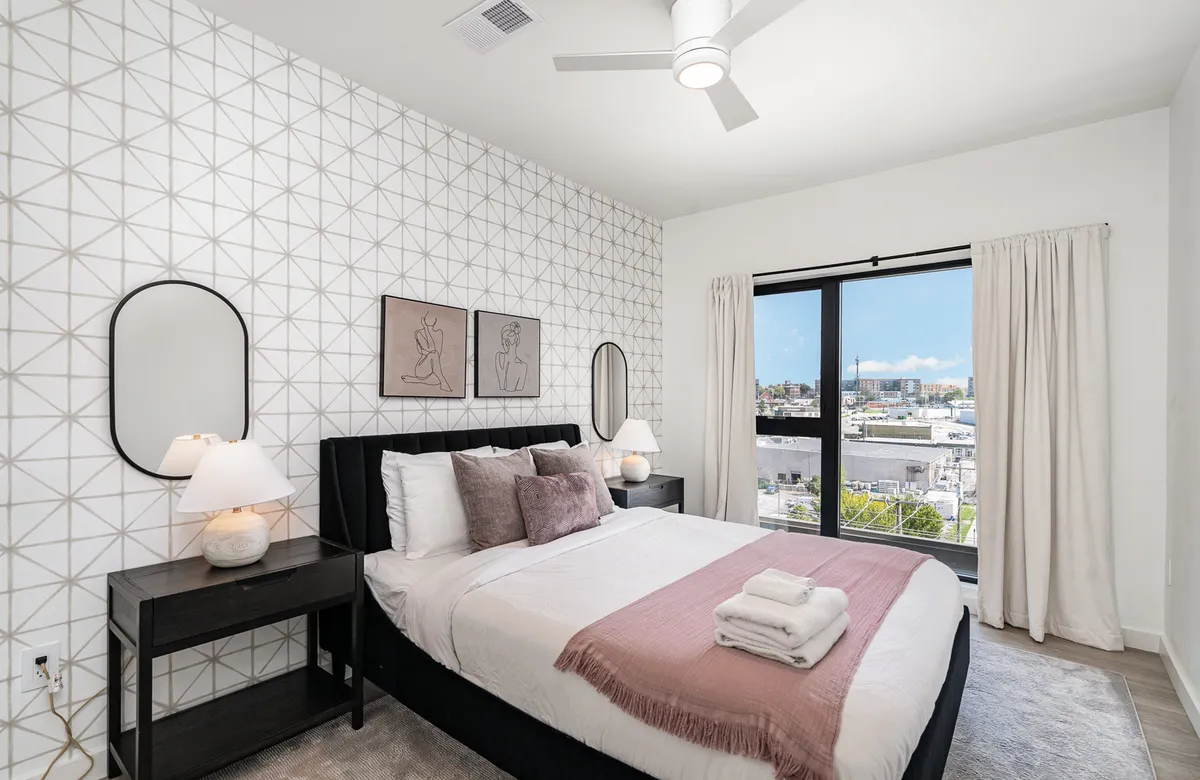 A tranquil bedroom with a bed dressed in modern linens, a large window letting in natural light, and a geometric patterned accent wall. The vibe is peaceful, with plenty of space to relax and settle in.