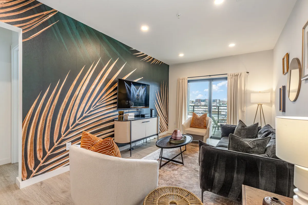 The image showcases a chic, sunlit living area featuring a vibrant tropical mural, plush modern sofas arranged for conversation and relaxation, a large flat-screen TV ideal for movie nights, and glass doors opening to a balcony with city and sunset views. The space radiates urban style and comfort, inviting guests to unwind or gather with friends against a colorful, Nashville-inspired backdrop.
