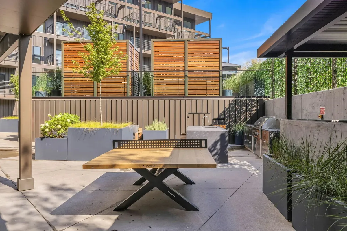 An outdoor communal area featuring a wooden table, green plants, wooden privacy screens, and a well-equipped grill setup, inviting guests to enjoy outdoor dining and barbecues in a casual, friendly atmosphere.
