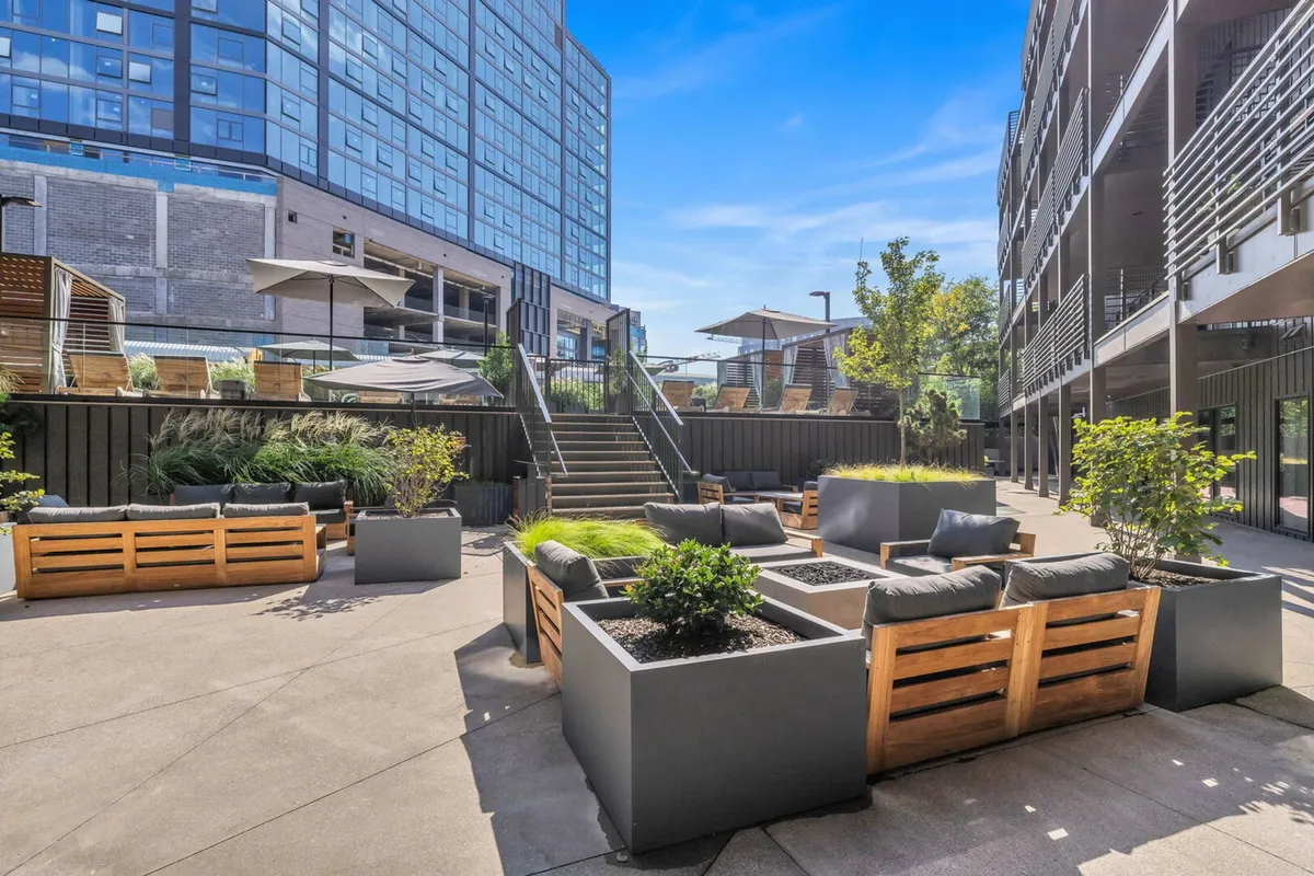 The image features the building's exterior entrance, surrounded by landscaped planters and stylish outdoor seating areas, with a staircase leading up to an upper level. The modern architectural lines of the property and surrounding city buildings are visible, and people are seen relaxing and socializing, hinting at the vibrant, urban environment.